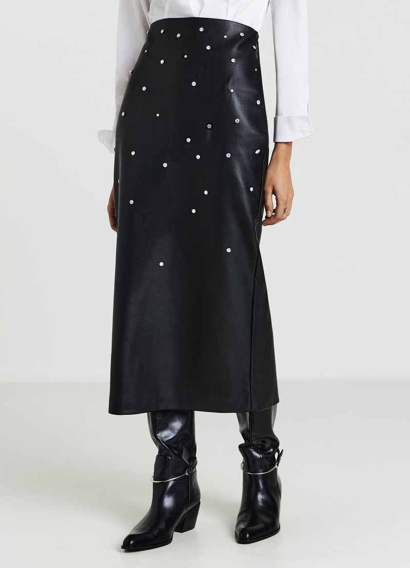 Jupe midi en cuir noir avec des clous décoratifs argentés, une fente latérale et une texture lisse, associée à des bottes à talons noires brillantes.