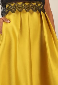 Gelbes Satin-Kleid mit einem schwarzen Spitzen-Oberteil. Der Rock hat Falten für zusätzliche Textur und Bewegung, was eine glatte Silhouette schafft.