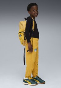 Gule sweatpants med broderet kattemotiv, parret med en sort langærmet skjorte og grønne sneakers, med en gul rygsæk ved siden af.