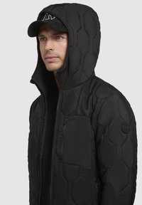 Schwarze Daunenjacke mit einem hexagonalen Steppmuster, ausgestattet mit einem hohen Kragen, einer verstellbaren Kapuze und einer mit Reißverschluss versehenen Brusttasche. Mit einem Logo-Patch akzentuiert.