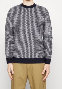 Laivastonsininen ja harmaa neule, jossa on teksturoitu aaltokuvio, resorikaulus ja -hihansuut, yhdistetty khakiväreihin housuihin. Materiaali vaikuttaa pehmeältä ja keskivahvalta.