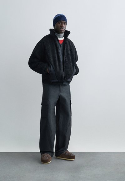 Crna flis jakna na patentni zatvarač, tamne cargo hlače s džepovima, smeđe cipele na navlačenje s uzorkom i plava pletena kapa. Teksturirani, topao izgled.