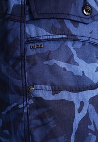 Blå kamouflage skjorta med en strukturerad tyg, framficka med tryckknapp och en liten metallisk "RAW"-logotyp på bröstet.