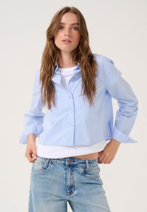 Light blue cropped button-up shirt made of cotton, featuring long sleeves with rolled cuffs and a single chest pocket.