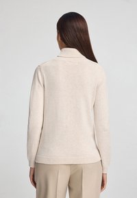 Maglione beige a maglia con colletto alto, maniche lunghe e orlo a coste. Texture morbida, design aderente, visto da dietro su uno sfondo chiaro.