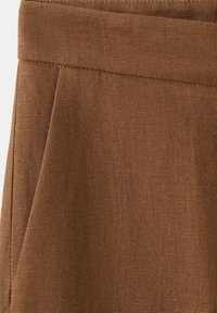 Smeđa teksturirana tkanina izbliza koja prikazuje pojas i šav bočnog džepa na odjevnom predmetu, vjerojatno od lana ili sličnog materijala.