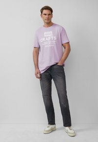 Lichtpaarse T-shirt met witte tekst, gecombineerd met donkergrijze jeans. Sneakers zijn crème met bruine accenten. Minimalistisch ontwerp, relaxed fit.