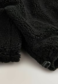 Zwart fleece stof met een gestructureerd, pluizig oppervlak; heeft een toggle en elastisch koord voor aanpassingsmogelijkheden.