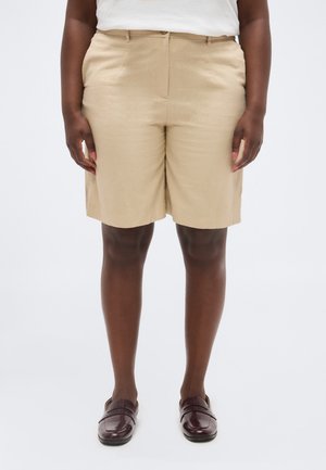 Person trägt beige knielange Shorts, weißes Oberteil und dunkelbraune Slipper, steht mit entspannt an den Seiten hängenden Händen.