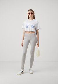Cropped white t-shirt with blue shell graphic, gray leggings, and white sneakers. Model holding a textured, circular white bag.