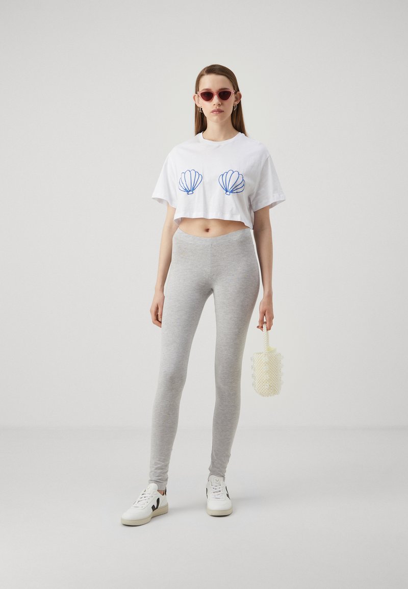 Cropped white t-shirt with blue shell graphic, gray leggings, and white sneakers. Model holding a textured, circular white bag.