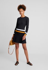 Melna adīta blūze ar garām piedurknēm un horizontālām svītrām baltā, oranžā un dzeltenā krāsā vidukļa daļā; apvienota ar melnu platmalu svārku un melnām sandalēm.