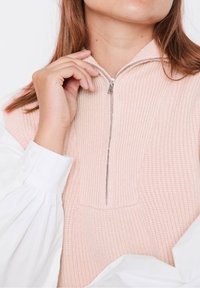 Hellrosa, strukturiertes Sweatshirt mit einem halben Reißverschluss am Kragen. Die weißen Manschetten des Shirts sind sichtbar und erzeugen einen geschichteten Effekt. Dezente Akzent-Reißverschlussdetails.