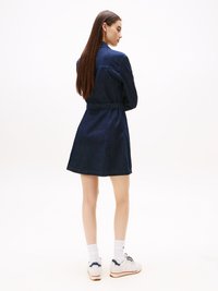 Denimdress, tummansininen, nappisuljettu etuosa, vyötärösolmu, levenevä helma, pitkät hihat, yhdistetty valkoisten tenniskenkien ja sukkien kanssa, nähtynä takaa.