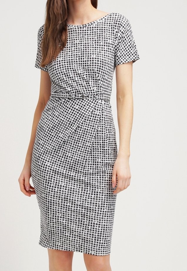 Femme portant une robe à carreaux noirs et blancs, à manches courtes et longueur genou, avec une ceinture fine à la taille.
