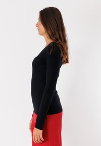 Top a maniche lunghe nero a coste con scollatura a V, silhouette aderente, abbinato a una gonna rossa. Il materiale appare morbido e strutturato.