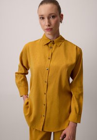 Hořčičně žlutá košile s límcem na knoflíky, dlouhými rukávy a dekorativními kovovými tečkami; měkká tkanina s volným střihem.