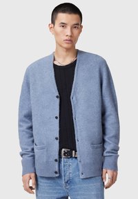 Cardigan azzurro chiaro realizzato in tessuto morbido, design aperto con cinque bottoni neri, due tasche frontali e polsini a coste. Indossato sopra una maglietta nera.