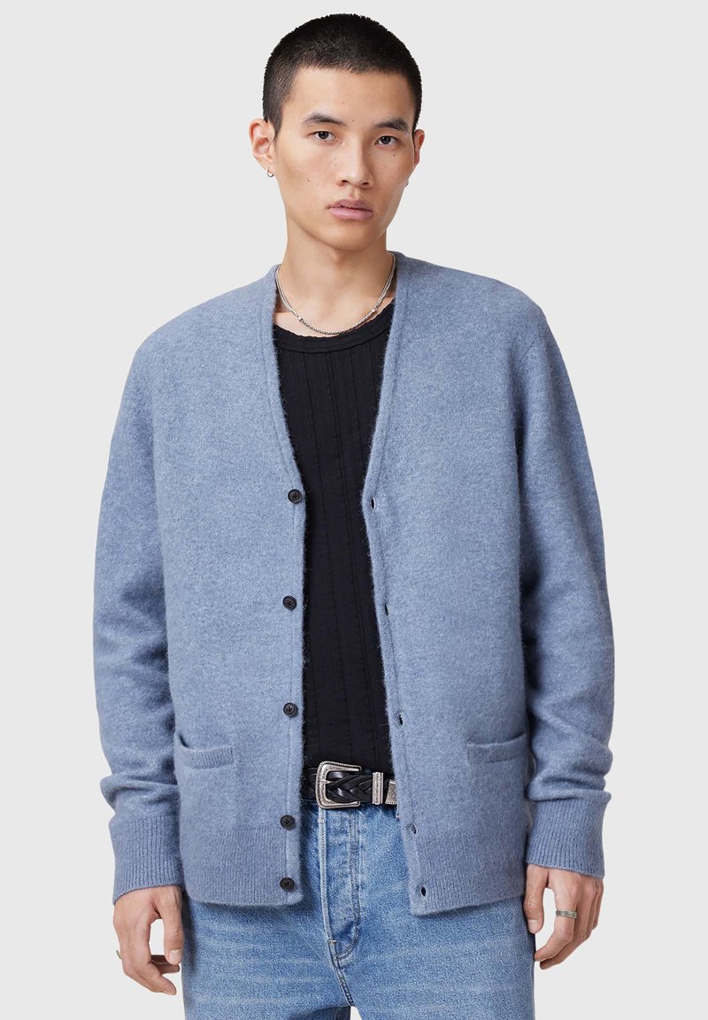Cardigan azzurro chiaro realizzato in tessuto morbido, design aperto con cinque bottoni neri, due tasche frontali e polsini a coste. Indossato sopra una maglietta nera.