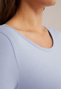 Närbild av en kvinna i en ljusblå topp med rundad halsringning, som visar hennes nacke, axel och övre bröstområde.