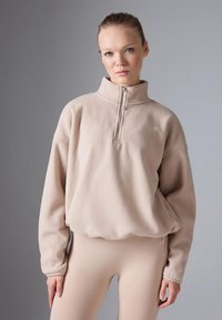 Beige fleece trui met een halve rits bij de kraag, losse pasvorm en geribbelde boorden. Gladde textuur, minimale branding aan de voorkant.