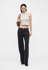 Femme tatouée portant un crop top sans manches rayé, un jean large noir, des bottes noires et un sac à bandoulière vert.