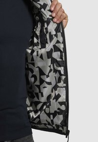Schwarzes und graues gemustertes Innenfutter einer Jacke mit glänzender Oberfläche, Reißverschluss und einer Hand, die die Jacke offen hält.
