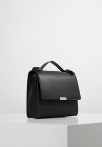 Černá kožená kabelka s výrazným designem, plochým vrškem a zlatým zapínáním. Má rukojeť a hladkou texturu.