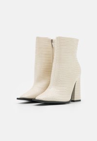 Białe botki sięgające kostki z teksturowanym wzorem krokodyla, spiczastym noskiem i słupkowym obcasem. Posiadają boczny zamek błyskawiczny dla łatwego zakładania.