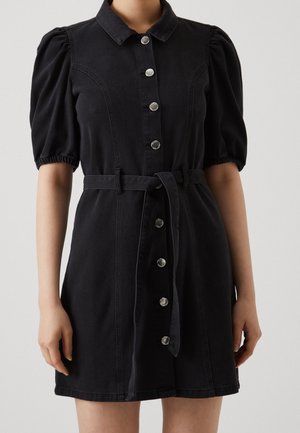 Zwarte spijkerjurk met korte pofmouwtjes, knoopsluiting aan de voorkant en een ceintuur in de taille. Voorzien van zilverkleurige knopen en een getailleerd silhouet.
