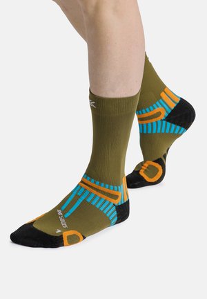 Benen met olijfgroene sportsokken met blauwe, oranje en zwarte patronen, staand op de tenen tegen een witte achtergrond.