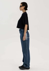 Geknipte zwarte t-shirt met brede mouwen, gecombineerd met losse blauwe jeans met een wit logo op de pocket, gedragen met zwarte sneakers.
