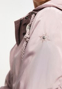 Veste rose clair avec une cordon à tressage, une fermeture à pression et à fermeture éclair, et un détail brodé en forme d'étoile sur l'épaule gauche.