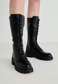 Black leather knee-high boots with a lace-up front, side zipper, chunky rubber sole, and textured detailing on the upper.