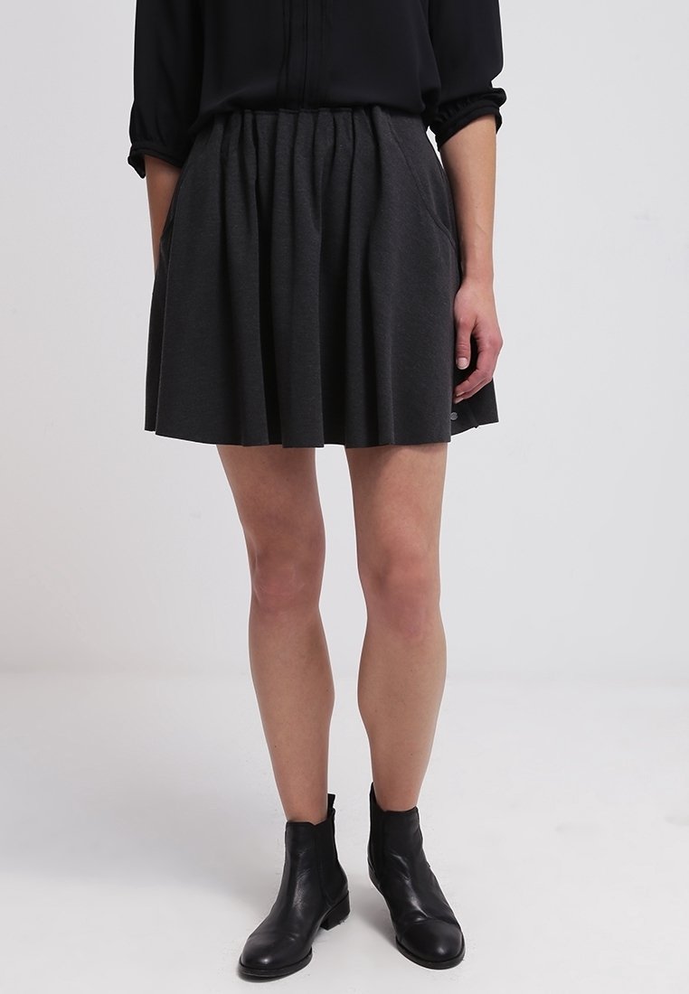 Femme portant une jupe plissée noire, un top noir à manches longues rentré, et des bottines noires, debout devant un fond clair uni.