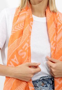 Personne ajustant une écharpe orange vif à motifs sur un t-shirt blanc et un jean bleu délavé, mettant l'accent sur les mains et le haut du torse.
