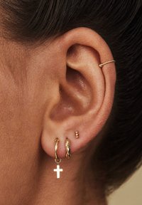 Gold hoop earrings, one with a cross charm, another twisted design, and a minimalist stud. Close-up on textured skin of the ear.