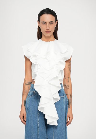 Femme aux cheveux noirs raides portant un haut en ruffles blanc à couches et un jean large bleu, montrant des tatouages sur les bras, se tenant devant un fond uni.