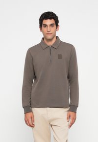 Mann iført en brun langermet polo med en kort glidelås og beige bukser, stående mot en hvit bakgrunn.