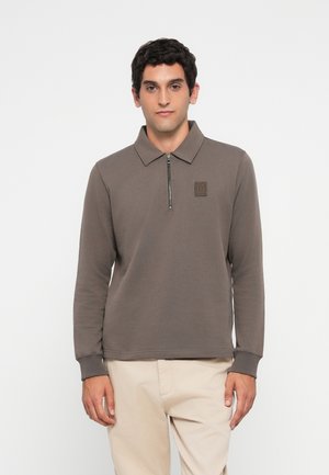 Man wearing a brown long-sleeve polo shirt with a short zipper and beige pants, standing against a white background.