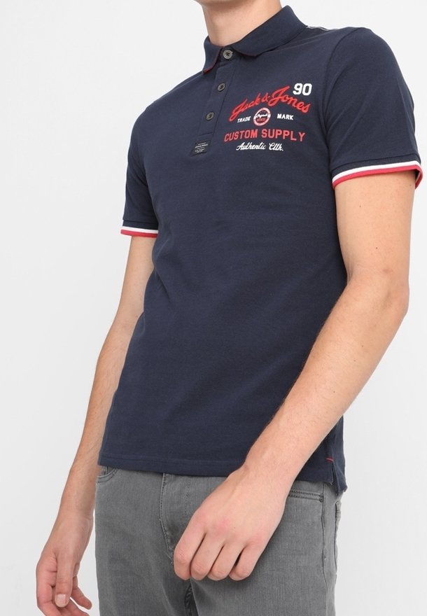 Homme portant un polo bleu marine avec des bordures rouge et blanche aux manches et un texte rouge sur la poitrine gauche, associé à un pantalon gris.