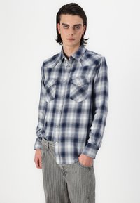 Camicia di flanella a scacchi blu e bianchi con un design a bottoni, due tasche sul petto e una consistenza liscia. Abbinata a pantaloni a righe grigi.