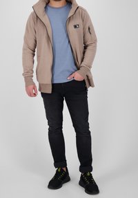 Felpa beige con zip e grande colletto, accompagnata da una t-shirt azzurra chiara, jeans neri e scarpe nere con dettagli verdi.