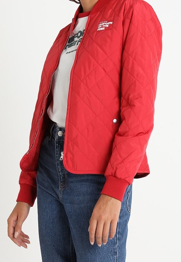 Personne portant une veste matelassée rouge sur un t-shirt imprimé blanc et un jean bleu, se tenant devant un fond blanc uni.