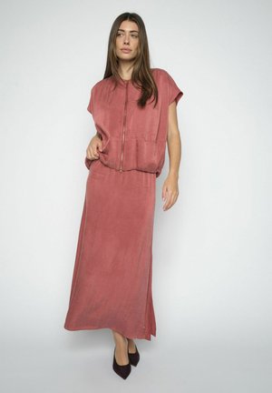 Kobieta ubrana w luźną, różowo-pudrową bluzę na suwak z krótkimi rękawami oraz dopasowaną długą, zwiewną spódnicę, do tego czarne, spiczaste buty.