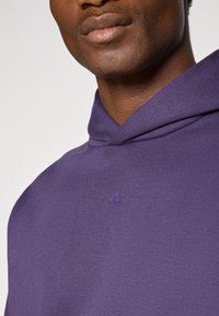 Paarse hoodie gemaakt van zachte stof, met een kangoeroezak en een subtiel geperst logo op de borst. Soepele textuur, losse pasvorm.