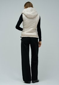 Ženska nosi svetlo bež č quilted bezrokavnik s kapuco, crno dolgi-sleeve top, široke črne hlače in črne čevlje s peto, obrnjena stran.