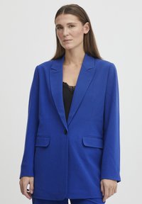 Královsky modrý blazer s klopami, jednoduchým zapínáním na knoflík a bočními kapsami. Hladká látka, volný střih doplněný černým krajkovým topem.
