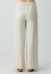 Vrouw in losse, hooggehaalde beige broek met elastische tailleband en zwarte open teen sandalen, staande tegen een lichte achtergrond.