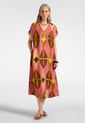 Mujer con cabello gris que lleva un vestido midi rosa con grandes formas geométricas marrones y amarillas, zapatos marrones, joyas de oro y está de pie sobre un fondo blanco.